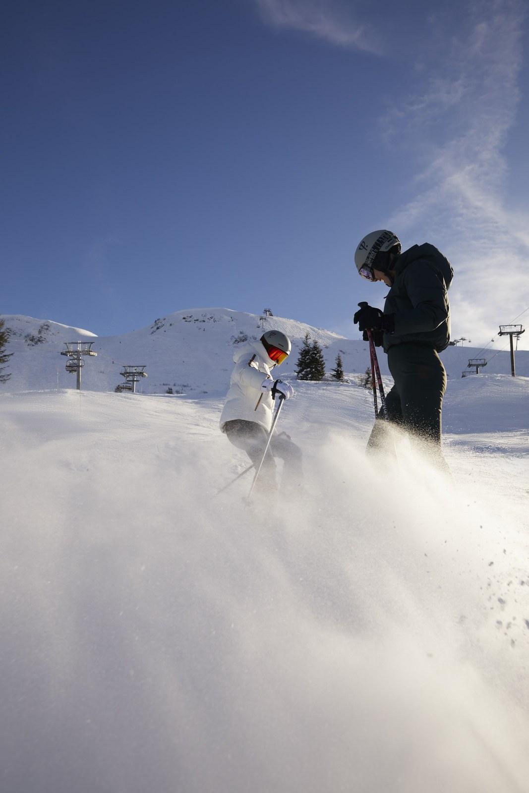 Two skiers in flying snow at Grand Massif with ski lifts and mountains behind